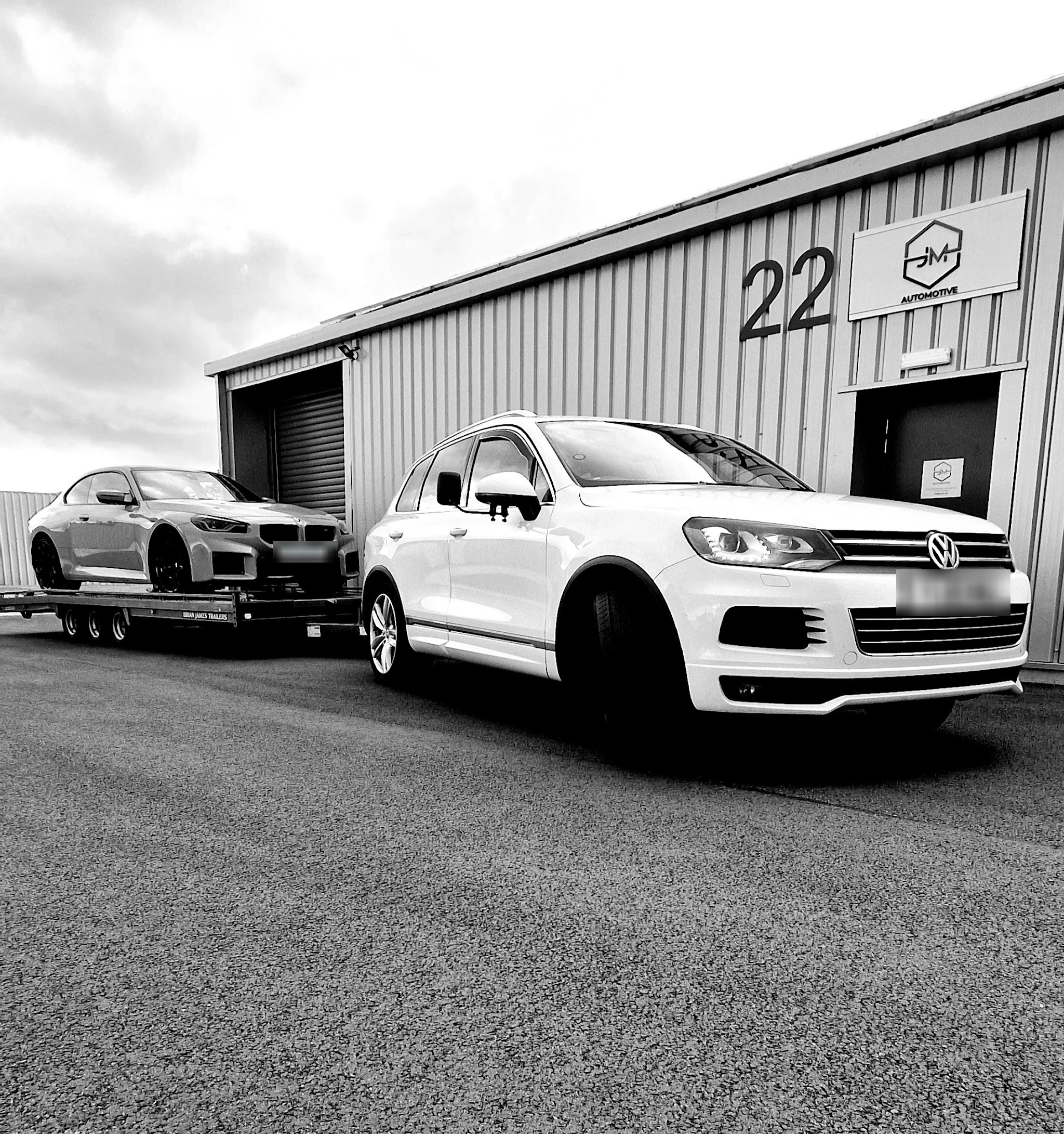 Black and white photo of JMM Automotive's unit 22 with the JM sign, a white Volkswagen Touareg and a car on a transporter trailer outside