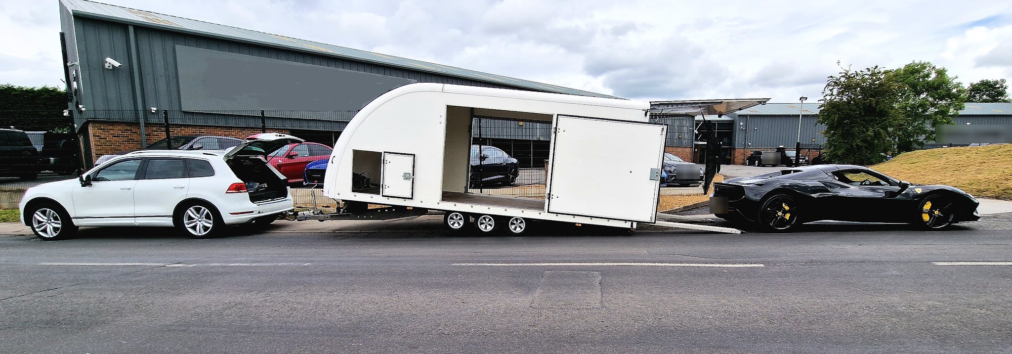 A white car loaded into an enclosed car transporter trailer on the trading estate, with a black sports car alongside
